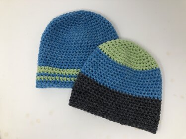 Zwei gehäkelte Mützen in Blau, Hellgrün und Dunkelgrau nebeneinander auf hellem Untergrund
