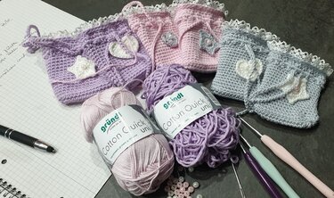 Drei gehäkelte Pastellbeutel mit Garnknäueln, Häkelnadeln und Perlen auf einer Arbeitsfläche.