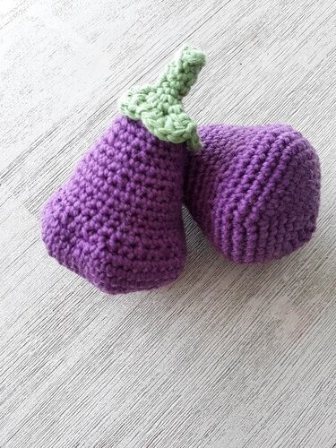 Zwei lila Amigurumi-Auberginen mit grünem Blattstiel nebeneinander auf hellem Untergrund