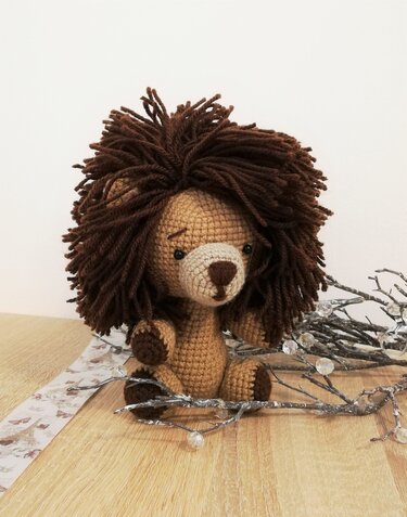 Gehäkelter Amigurumi-Löwe mit zotteliger brauner Mähne, sitzend auf einem Holztisch neben Dekozweigen.