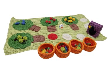 Praktisches Häkel-Set mit vier Körbchen, gehäkelten Früchten, Baum-Motiven auf grüner Spielmatte, einem Würfel und einer lila Vogel-Spielfigur