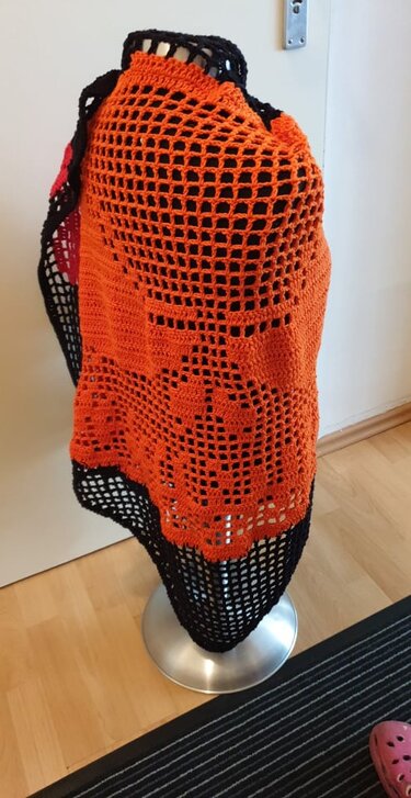 Orange-schwarzes gehäkeltes Tuch mit großem Netzmuster auf einem silbernen Ständer.