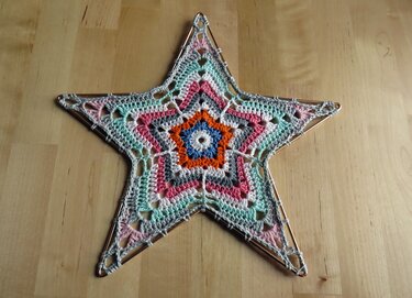 Fünfzackiger gehäkelter Stern in Pastellfarben auf Drahtrahmen auf hellem Holztisch.