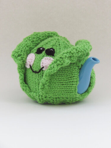 Blue-spouted teapot fitted with a green knitted cabbage tea cosy featuring a smiling face and folded leaf panels