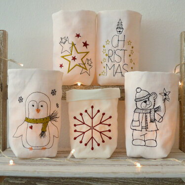 Fünf bestickte Hüllen für LED-Tischlichter mit Pinguin, Bär, Sternen, Schneeflocke und Schriftzug CHRISTMAS auf Holzregal mit Lichterkette.