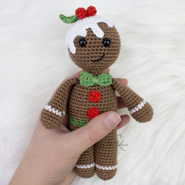 Gehäkeltes Lebkuchenmännchen-Amigurumi mit Zuckerguss-Optik, Stechpalme, grüner Fliege und roten Knöpfen, in einer Hand vor weißem Plüsch.