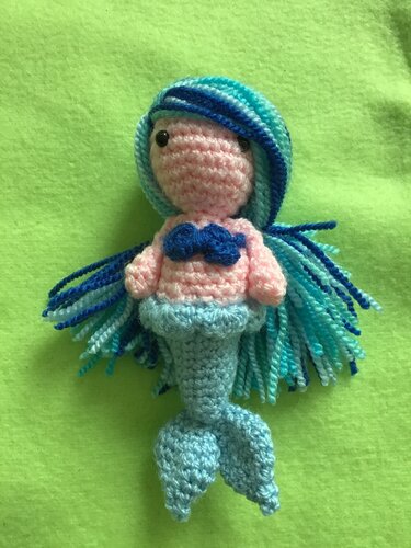Gehäkelte Amigurumi-Meerjungfrau mit rosa Körper, blauem Muschel-Oberteil, hellblauer Schwanzflosse und blau-türkisem Garnhaar auf grünem Hintergrund.
