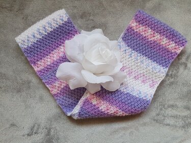 Zwei gestrickte Kinder-Nackenwärmer in Pastellfarben mit weißer Stoffrose auf hellem Untergrund.