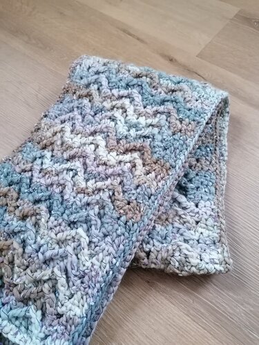 Gefalteter, dicker Häkelschal mit Strukturmuster und mehrfarbigem Garn in Blau-, Grau- und Lilatönen auf hellem Holzboden.