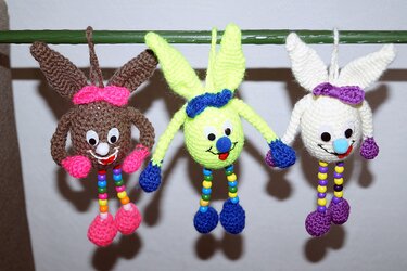 Drei gehäkelte Hasen (braun, neongrün, weiß) mit Kulleraugen und Perlenbeinen an einer grünen Stange hängend
