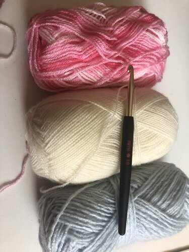 Drei zarte Wollknäuel in Rosa, Creme und Hellblau/Grau mit Häkelnadel.