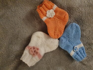 Drei kleine gestrickte Neugeborenen-Socken (orange, weiß, blau) mit Rippbündchen und Applikationen