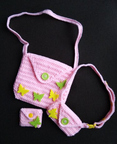 Rosa gehäkelte Umhängetasche mit Klappe, grünem Knopf und gelb-grünen Schmetterlings-Applikationen, dazu kleine passende Täschchen auf schwarzem Hintergrund