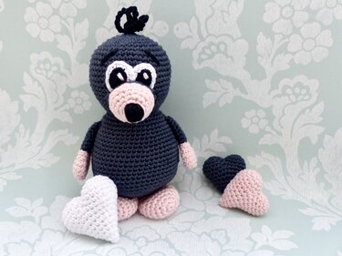 Gehäkelte dunkelgraue Amigurumi-Maulwurf-Figur mit rosa Schnauze, rosa Pfoten und drei gehäkelten Herzen in Weiß, Rosa und Dunkelgrau