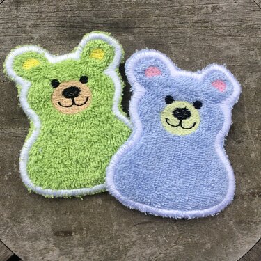 Zwei gestickte Bären-Applikationen in Frottee-Optik, grün und hellblau, mit weißen Konturen und bestickten Gesichtern auf Holzuntergrund