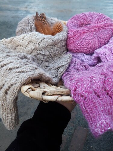 Hand hält Korb mit grauen und violetten gestrickten Armstulpen, Wollknäueln und dekorativen Federn.