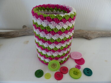 Zylindrischer gehäkelter Utensilo in Pink, Grün und Weiß mit Muschelmuster, davor verstreute bunte Knöpfe auf weißer Oberfläche.