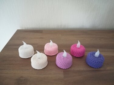 Sechs gehäkelte Hüllen in Weiß, Creme, Rosa, Pink, Flieder und Violett über batteriebetriebenen LED-Teelichtern auf einem Holztisch