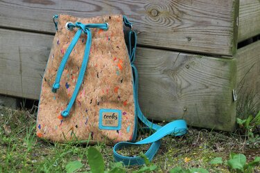 Kleine Beuteltasche in Korkoptik mit bunten Sprenkeln, türkisfarbener Kordel und langem Träger, steht vor einer Holzverkleidung
