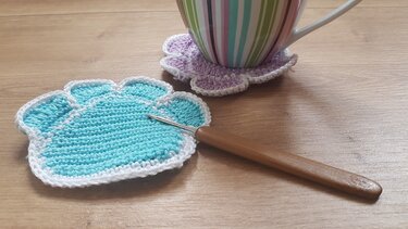 Türkis und lila gehäkelte Pfoten-Untersetzer mit weißer Kante und Häkelnadel auf Holztisch