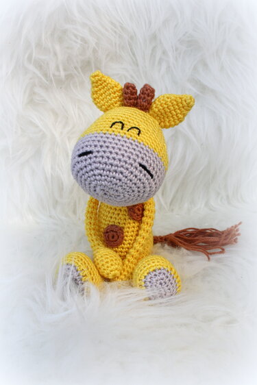 Gehäkelte gelbe Amigurumi-Giraffe sitzend mit hellgrauem Maul und braunem Schwanz