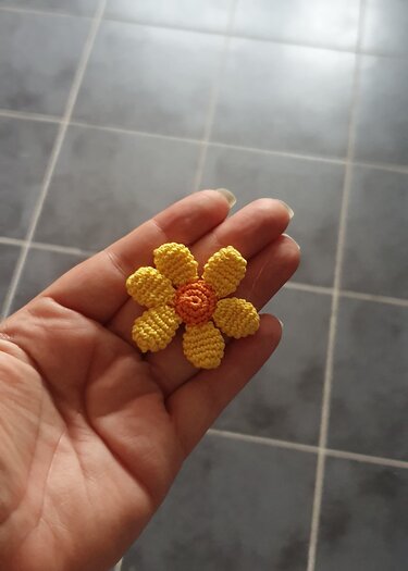 Hand präsentiert eine kleine, schnell gehäkelte Blume mit sechs gelben Blütenblättern und orangefarbenem Zentrum