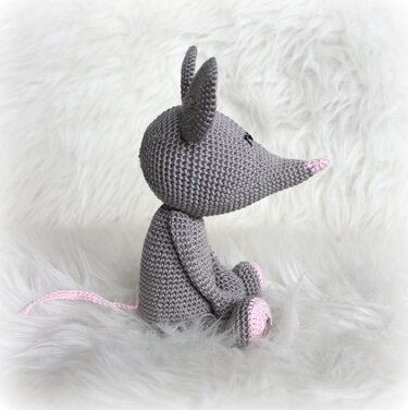 Gehäkelte graue Amigurumi‑Maus mit rosa Nase, Ohren und Pfoten auf weißem, flauschigem Untergrund, seitlich gesehen.