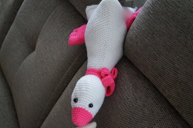 Nahaufnahme einer weißen gehäkelten Amigurumi-Gans mit pinkem Schnabel, pinken Flügeln und kleinen schwarzen Augen auf braunem Sofa