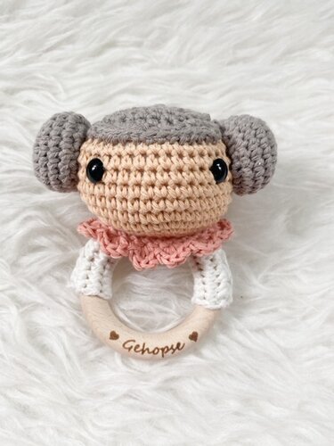 Gehäkelte Babyrassel mit hellem Gesicht, grauen Haarknoten, rosafarbenem Kragen und Holzring auf weißem, flauschigem Untergrund.