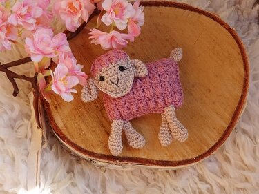 Gehäkeltes Amigurumi-Lamm mit altrosafarbenem Körper auf einer Holzscheibe neben rosa Blüten