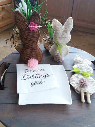 Zwei gehäkelte Hasen in Braun und Creme mit Bommel-Schwänzchen und grünen Schleifen neben Teller und Serviette 'Für meine Lieblingsgäste'.