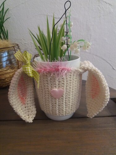 Weiße Tasse mit cremefarbener Häkelhülle, seitlichen Hasenohren, rosa Herzknopf und grün-kariertem Schleifenband