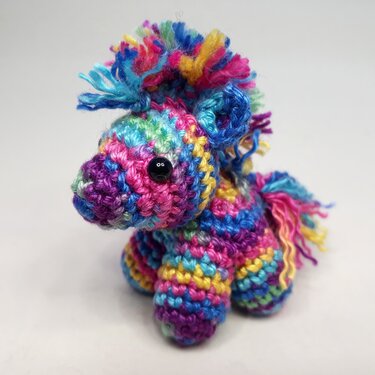 Gehäkeltes Amigurumi-Pony in Regenbogenfarben mit zotteliger Mähne und schwarzen Augen vor hellem Hintergrund.
