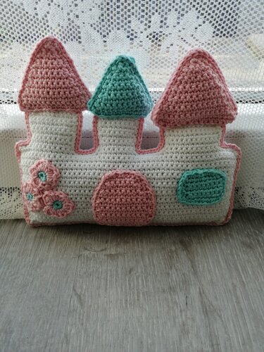 Gehäkeltes Schloss-Kissen in Pastellfarben vor weißer Gardine auf Holzboden
