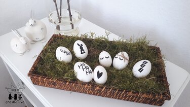 Korb mit Moos und weißen Eiern, verziert mit schwarzen Plottermotiven und Deko-Hasen im Hintergrund