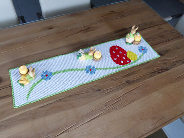 Weißer gehäkelter Tischläufer mit rotem Pixel-Ei, blauen Blumen und grüner Bordüre, dekoriert mit Hasen-Kerzenhaltern auf einem Holztisch