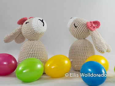 Zwei Amigurumi-Hasen in hellem Beige mit rosa Schleifen, sitzend neben bunten Ostereiern.