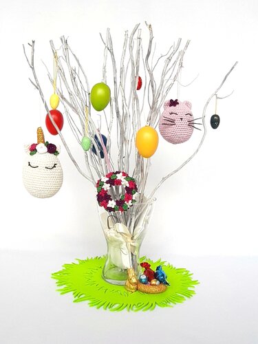 Vase mit weiß lackierten Zweigen, daran zahlreiche gehäkelte Osteranhänger (Einhorn, Katzenkopf, Blumenkranz) in Pastell- und kräftigen Farben; grüner Filzuntersetzer mit kleinen Schokoladenfiguren.