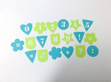 Gestanzte Zahlen 0–9 und Symbole wie +, =, ?, ! in türkisen und limettengrünen Formen auf weißem Hintergrund.