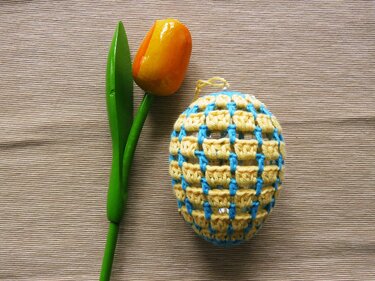 Gehäkeltes gelb-hellblaues Oster-Ei mit Gittermuster neben einer orange-gelben Holz-Tulpe auf beigem Stoff.