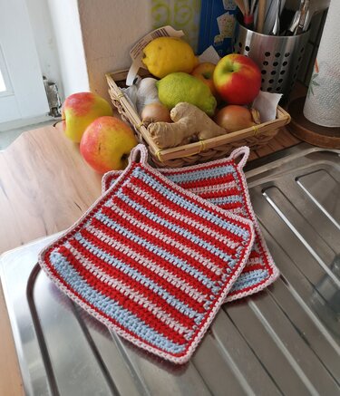 Zwei handgehäkelte Topflappen mit horizontalen roten, hellblauen und weißen Streifen, rosa Rand und Aufhängeschlaufen auf einer Küchenspüle neben einer Obstschale.