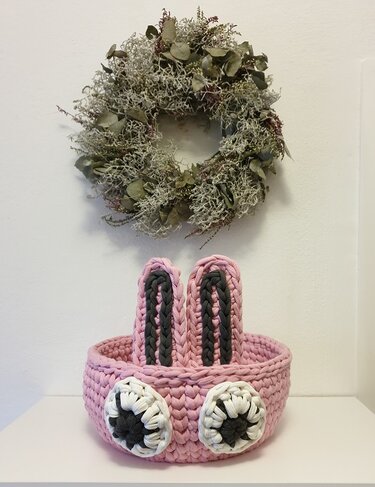 Hellrosa gehäkelter Korb mit zwei aufrechten Hasenohren und weißen runden Applikationen vor heller Wand