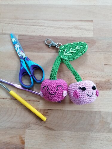 Zwei gehäkelte Amigurumi‑Kirschen mit lächelnden Gesichtern als Schlüsselanhänger neben Häkelnadel und Schere