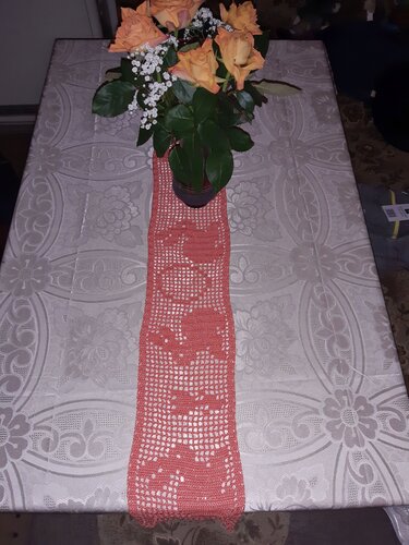 Lachsfarbener gehäkelter Tischläufer mit filigranem Muster, darauf ein Blumenstrauß aus orangen Rosen.