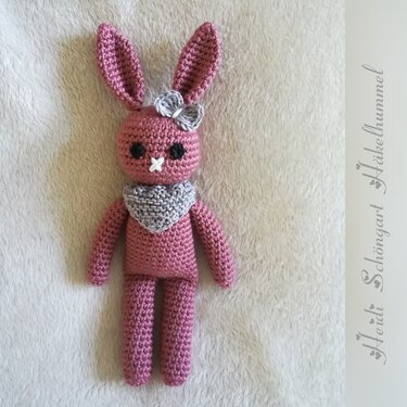 Rosa gehäkelter Amigurumi-Hase mit grauer Schleife am Ohr und grauem Schal auf hellem, flauschigem Untergrund.
