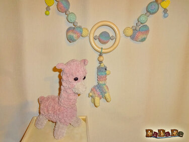 Rosa, flauschig gehäkeltes Lama-Plüschtier neben einem vielfarbigen Alpaka-Hängespielzeug an einem Holzring mit gehäkelten Perlen.