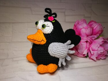 Schwarzer gehäkelter Amigurumi-Vogel mit weißer Gesichtsmaske, grünen Augen, orangefarbenem Schnabel und rosa Rose auf dem Kopf.