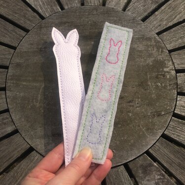 Hand hält zwei ITH-Lesezeichen mit Hasenkonturen: ein längliches Filzstück mit drei gestickten Hasen und ein geformtes, helles Lesezeichen aus Kunstleder