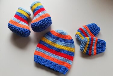 Nahaufnahme eines gestrickten Baby-Sets mit gestreifter Mütze, Schühchen und Fäustlingen in Blau, Gelb, Rot und Orange auf hellem Hintergrund.