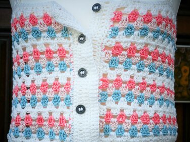 Nahaufnahme einer handgehäkelten Damenweste im Granny-Muster in Weiß, Rosa und Hellblau mit sichtbarer Knopfleiste